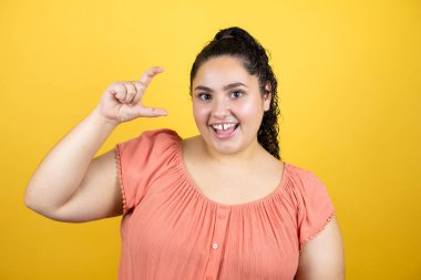 Sarı arka planda izole edilmiş kıvırcık saçlı genç bir kadın gülümsüyor ve kendinden emin bir şekilde el hareketleriyle küçük beden işareti yapıyor parmakları bakıyor ve kameraya bakıyor. Ölçü kavramı.