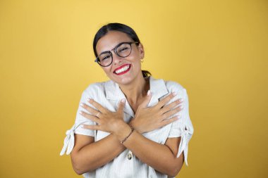 Sarı arka planda güzel bir kadın kendine sarılıyor mutlu ve pozitif, kendinden emin gülümsüyor.