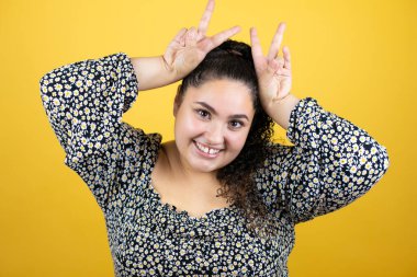 Sarı arka planda kıvırcık saçlı genç bir kadın komik ve çılgın pozlar veriyor parmakları tavşan kulaklı, neşeli gülümsüyor.