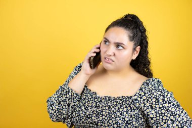 Sarı arka plan üzerinde kıvırcık saçlı genç ve güzel bir kadın telefonda şaşkın bir ifadeyle konuşuyor.