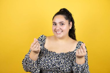 Kıvırcık saçlı, soyutlanmış sarı arka planda genç bir kadın. Elleriyle para hareketi yapıyor. Maaş ödemesi istiyor.