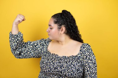 Sarı arka planda kıvırcık saçlı güzel bir kadın gururla gülümseyen kol kaslarını gösteriyor.