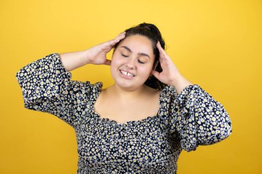 Kıvırcık saçlı genç bir kadın izole edilmiş sarı arka planda rahatlıyor ve esniyor, kolları ve elleri başının arkasında mutlu bir şekilde gülümsüyor.