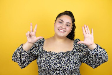 Sarı arka planda kıvırcık saçlı genç bir kadın sekiz numaralı parmaklarıyla gülümseyerek kendinden emin ve mutlu olduğunu gösteriyor.