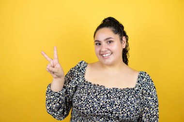 Sarı arka planda kıvırcık saçlı genç bir kadın iki numaralı parmaklarıyla gülümseyerek kendinden emin ve mutlu olduğunu gösteriyor.