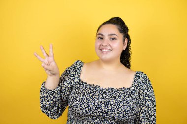 Sarı arka planda kıvırcık saçlı genç bir kadın üç numaralı parmaklarıyla gülümseyerek kendinden emin ve mutlu olduğunu gösteriyor.