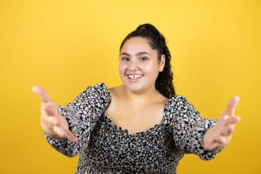 Sarı arka planda kıvırcık saçları olan genç bir kadın kameraya sarılıp gülümsüyor.