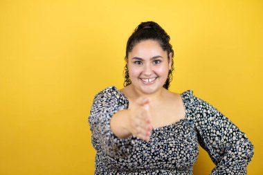 Sarı arka plan üzerinde kıvırcık saçları olan genç ve güzel bir kadın. Dostça bir selamlama ve hoş geldin hediyesi olarak gülümsüyor.