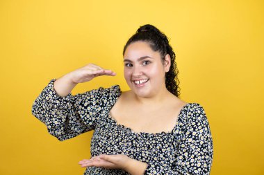 İzole edilmiş sarı arka plan üzerinde kıvırcık saçlı genç ve güzel bir kadın. Ellerinde büyük ve büyük bir işaret var. Ölçü sembolü. Kameraya bakıp gülümsüyor. Ölçüm konsepti.