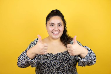 Sarı arka planda kıvırcık saçlı genç bir kadın gülümsüyor ve baş parmaklarıyla işaret veriyor.