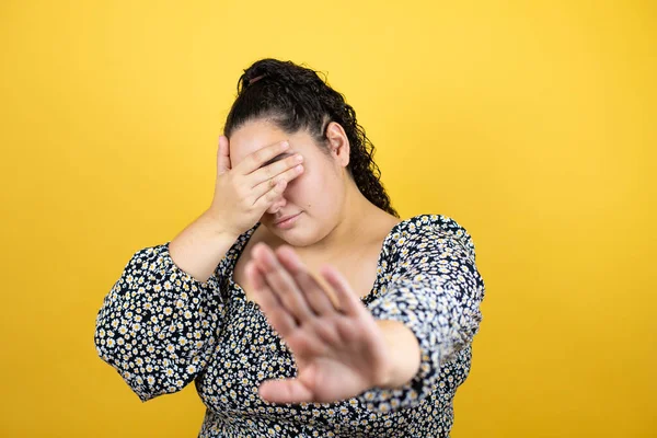 Young beautiful woman with curly hair over isolated yellow background covering eyes with hands and doing stop gesture with sad and fear expression. Embarrassed and negative concept.