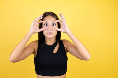 Güzel bir genç kız, izole edilmiş sarı arka planda parmaklarıyla gözlerini açmaya çalışıyor, uykulu ve yorgun bir şekilde sabah yorgunluğu çekiyor.