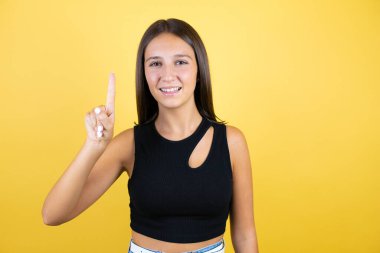 Güzel bir genç kız izole edilmiş sarı arka planda bir numaralı parmaklarıyla işaret ederken kendinden emin ve mutlu bir şekilde gülümsüyor.