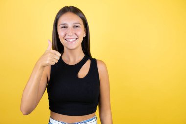 Güzel bir genç kız, izole edilmiş sarı arka planda gülümsüyor ve baş parmağıyla işaret veriyor.