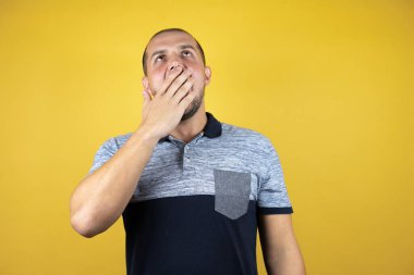 İzole edilmiş sarı arka planda dikilen Rus adam ağzıyla ağzını kapatmaktan sıkılmış. Huzursuz ve uykulu..