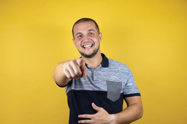 Rus adam sarı arka planda dikilip sana gülüyor, eli vücudunun üzerinde kamerayı işaret ediyor, utanç ifadesi.