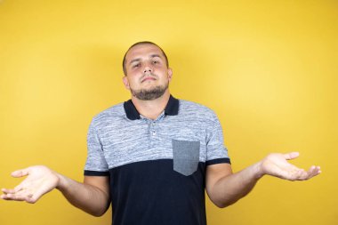 İzole edilmiş sarı arka planda dikilen, elleri ve kolları havada, kafası karışmış bir Rus adam.