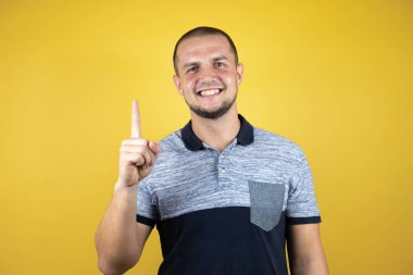 Rus adam sarı arka planda duruyor. Bir numarayı işaret ediyor ve gülümsüyor. Kendinden emin ve mutlu..