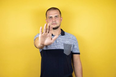 Korunmasız sarı arka planda dikilen Rus adam, eli açık, dur işareti yapıyor, ciddi ve kendinden emin bir ifadeyle, savunma hareketi yapıyor.