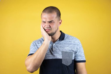 Rus adam, diş ağrısı ya da diş hastalığından dolayı sarartılmış sarı arka planda durup ağzıyla acı dolu bir ifadeye dokunuyor.