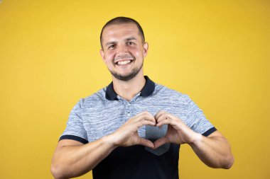 Korunmasız sarı arka planda dikilen Rus adam, ellerinde kalp sembolü ve şekli olan aşkla gülümsüyor. Romantik konsept.