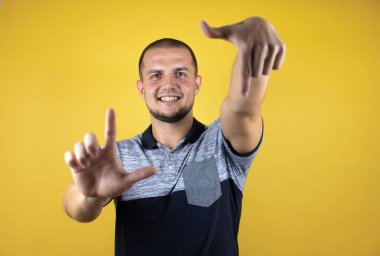 Rus adam sarı arka planda duruyor. El ve parmaklarıyla çerçeve yapıyor. Kamera açısı.