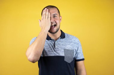 Rus adam sarı arka planda duruyor. Bir gözünü eliyle kapatıyor. Yüzünde kendine güvenen bir gülümseme ve sürpriz bir duygu var..