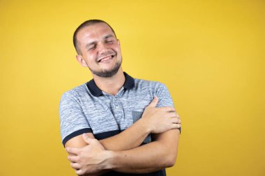 Rus adam sarı arka planda dikilip kendine sarılıyor mutlu ve pozitif, kendinden emin gülümsüyor.