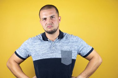 Sarı arka plan üzerinde duran Rus adam kuşkucu ve gergin, yüzünde tasvip etmeyen ifade belinde kollarla