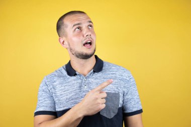 İzole edilmiş sarı arka planda dikilen Rus adam afallamış ve baş parmağıyla kenarı işaret ediyor..