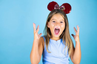 Fare kulağı takan genç ve güzel bir kız çocuğu. Mavi arka planda çılgın ve kızgın bir şekilde bağırıyor ve agresif bir ifade ve kolla bağırıyor.