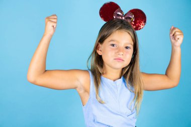 İzole edilmiş mavi arka planda fare kulağı takan güzel bir kız çocuğu gururla gülümseyen kol kaslarını gösteriyor.