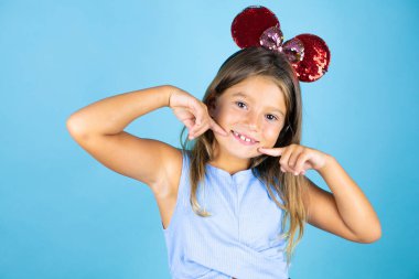 İzole edilmiş mavi arka planda fare kulağı takan genç ve güzel bir kız. Gülümsüyor ve parmaklarıyla ve ağzıyla işaret ediyor.