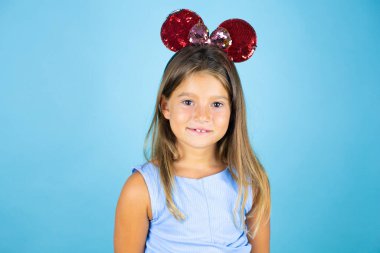 İzole edilmiş mavi arka planda fare kulağı takan güzel bir kız çocuğu kameraya bakıp gülümsüyor.