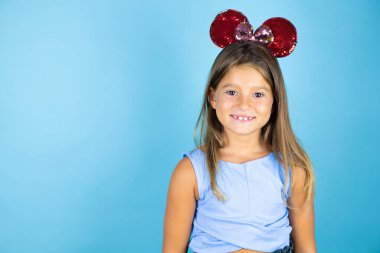 İzole edilmiş mavi arka planda fare kulağı takan güzel bir kız çocuğu kameraya bakıp gülümsüyor.