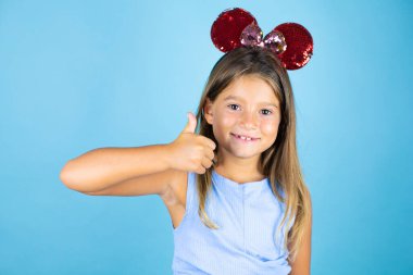 İzole edilmiş mavi arka planda fare kulağı takan güzel bir kız çocuğu gülümsüyor ve baş parmağıyla işaret veriyor.