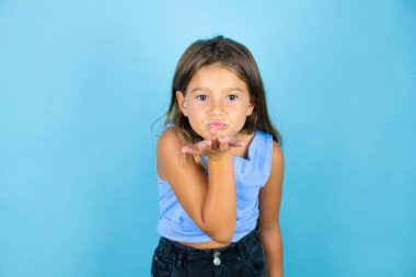 Genç, güzel bir kız çocuğu, izole edilmiş mavi arka planda kameraya bakıp havada bir öpücük savuruyor ve sevimli ve seksi oluyor. Aşk ifadesi.