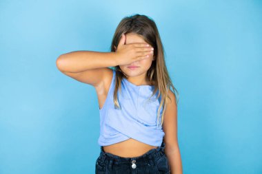 Genç, güzel bir kız çocuğu, soyutlanmış mavi arka planda ciddi ve elleriyle gözlerini kapatıyor.