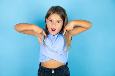 Genç, güzel bir kız çocuğu, izole edilmiş mavi arka planda, kendine güvenen, yüzünde bir gülümsemeyle, kendini gururlu ve mutlu bir şekilde gösteren..