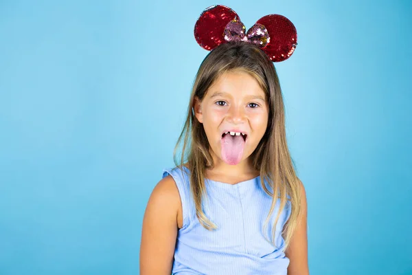 Young beautiful child girl wearing mouse ears over isolated blue background sticking out tongue very happy