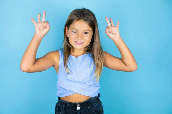 年轻美丽的小女孩在孤零零的蓝色背景上 用手指和微笑做手势 是个很好的象征 — 图库照片
