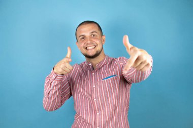 Rus iş adamı, mavi arka planda kırmızı gömlekli, mutlu ve komik yüzlü parmakları kameraya doğrultuyor. İyi enerji ve titreşimler.