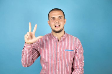 Rus iş adamı mavi arka planda kırmızı gömlek giyer ve üç numaralı parmaklarıyla gülümseyerek kendinden emin ve mutlu bir şekilde işaret eder..