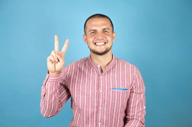 Rus iş adamı mavi arka planda kırmızı tişört giyer ve iki numaralı parmaklarıyla işaret ederken kendinden emin ve mutlu gülümserdi..