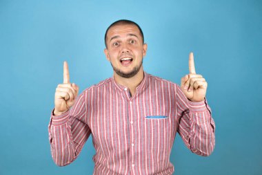 Rus iş adamı mavi arka planda şaşkın şaşkın yukarı bakar, parmaklarıyla işaret eder ve kollarını kaldırırdı.