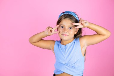 Güzel bir kız çocuğu pembe arka planda barış sembolü yapıyor. Yüzünde parmaklar, yüzünde neşeyle zafer işareti yapıyor.