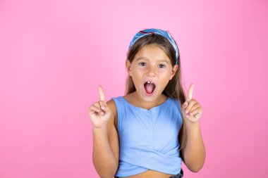 Güzel bir kız çocuğu pembe arka planda hayrete düşmüş ve şaşırmış bir şekilde kameraya bakıyor ve parmaklarıyla işaret ediyor ve kollarını kaldırıyor.