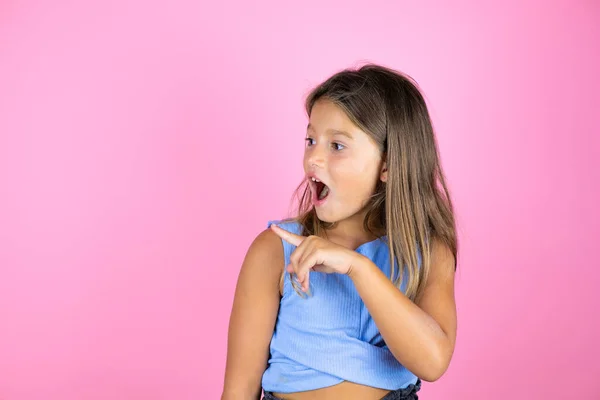 Genç, güzel bir kız çocuğu, soyutlanmış pembe bir arka planı var. Şaşırmış ve eli ve parmağı kenarı işaret ediyor.