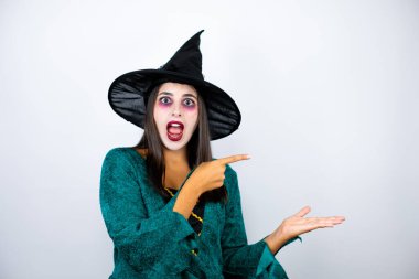 Cadı kostümü giymiş bir kadın beyaz arka planda şaşırmış ve elindeki bir şeyi gösterip işaret ediyor.
