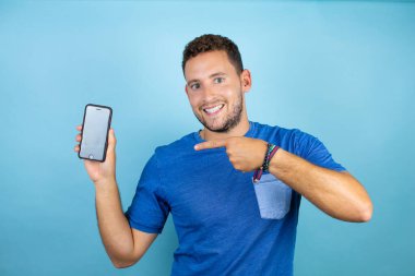 Genç ve yakışıklı bir adam mavi tişört giyip izole edilmiş mavi arka planda gülümsüyor ve telefonu işaret ediyor.